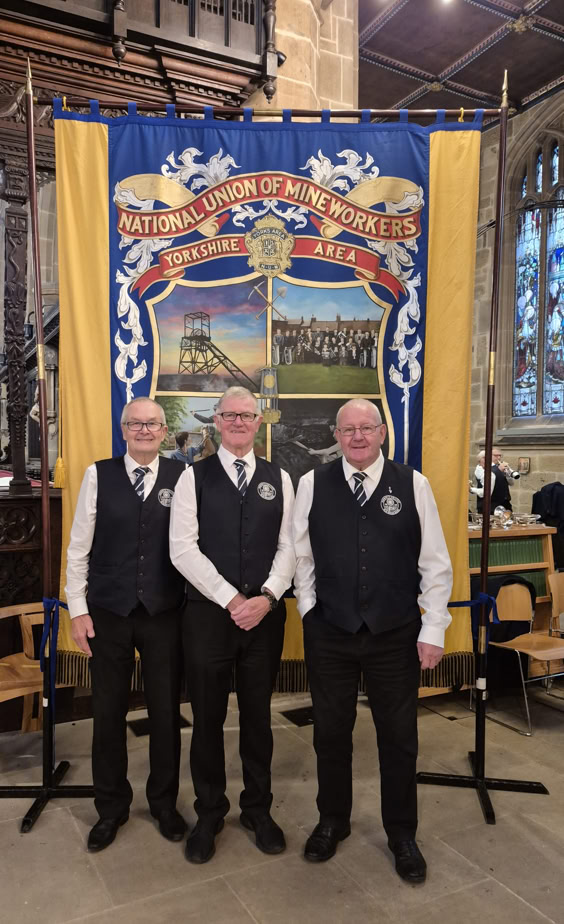 Image of Dodworth Colliery Brass Band members Keith Kellett, Mick Wood and Dave Swift stood in front of the National Union of Mineworkers Banner at Wakefield Cathedral.